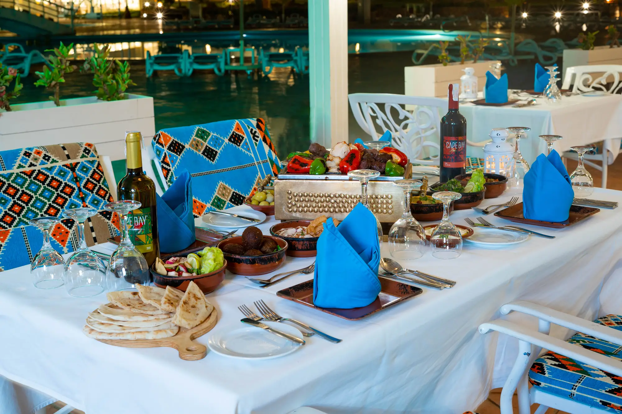Tavolo apparecchiato per cena all'aperto con piatti tipici, vino e tovaglioli blu vicino alla piscina illuminata