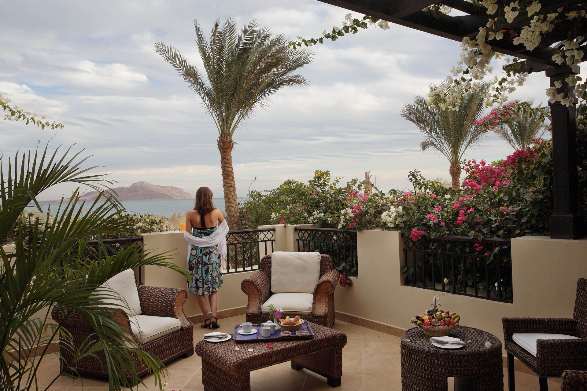 Terrazza privata con divani in vimini e donna che ammira la vista sul mare tra fiori e palme